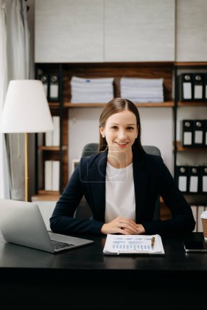 Téléchargez les photos : Femme d'affaires souriante en veste noire assise dans la table avec ordinateur portable, statistiques papier et sourire à la caméra - en image libre de droit