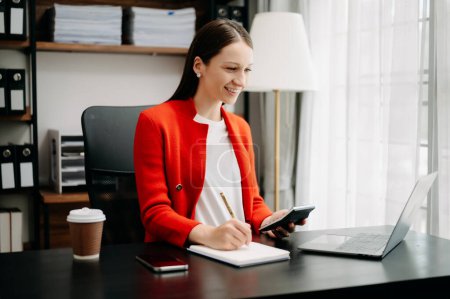 Téléchargez les photos : Femme d'affaires souriante en veste rouge écrit des notes tout en étant assis dans la table avec calculatrice, ordinateur portable et tasse à café - en image libre de droit