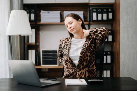 Foto de Mujer de negocios con exceso de trabajo en la mesa de la oficina, con chaqueta de leopardo, que sufre de dolor de espalda - Imagen libre de derechos