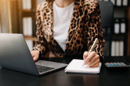 Photo for Cropped image of woman wearing leopard jacket, working at office desk with laptop and writing notes - Royalty Free Image