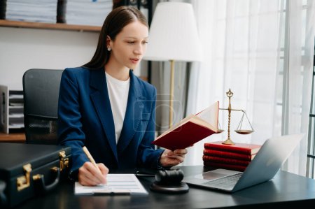 Téléchargez les photos : Belle femme caucasienne lecture livre de justice et de prendre des notes avec stylo avec travail dans le bureau à table avec ordinateur portable - en image libre de droit