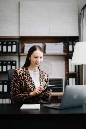 Téléchargez les photos : Femme d'affaires brune portant une veste léopard et assise à table avec un ordinateur portable. femme utilisant et tenant la calculatrice - en image libre de droit