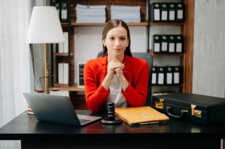 Foto de Mujer abogada caucásica con chaqueta roja y sentada en el escritorio de la oficina con laptop, martillo, estuche y carpeta, - Imagen libre de derechos