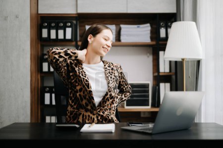 Foto de Mujer de negocios con exceso de trabajo en la mesa de la oficina, con chaqueta de leopardo, que sufre de dolor de espalda - Imagen libre de derechos