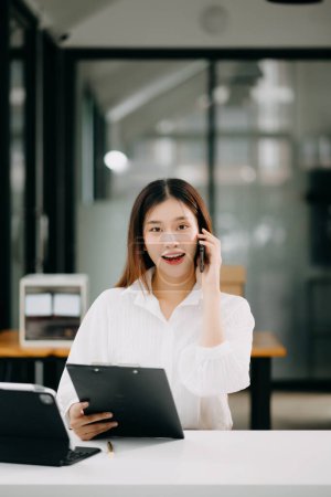 Photo for Young beautiful Asian woman talking on smartphone while sitting at the working white table office - Royalty Free Image