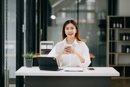 Photo for Young beautiful Asian woman drinking coffee while sitting at the working white table office - Royalty Free Image
