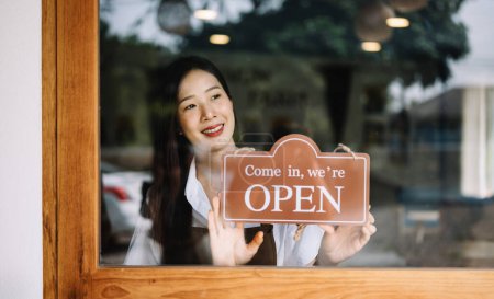 Foto de Dueño de una pequeña empresa. Mujer asiática mostrando un cartel de bienvenida frente a una cafetería. Mujer camarera, propietaria de coffeeshop. - Imagen libre de derechos