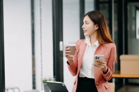 Photo for Confident Asian woman with a smile standing holding coffee and smartphone at the modern office - Royalty Free Image