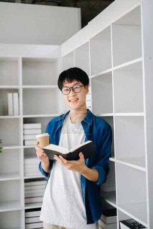 Téléchargez les photos : Étudiant mâle debout avec livre et café dans la bibliothèque du collège - en image libre de droit