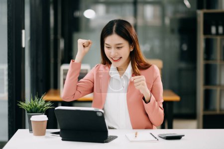 Photo for Young Asian businesswoman feeling excited about her professional success. Joyful woman making winner's gesture in office - Royalty Free Image