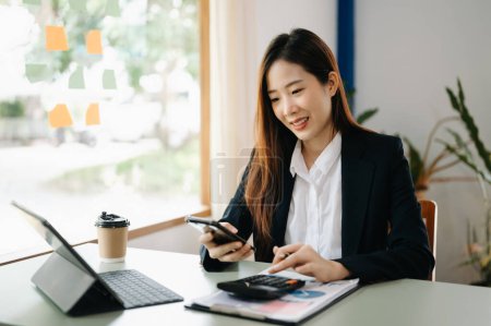 Foto de Mujer de negocios asiática que trabaja en la oficina del banco con calculadora. mesa con portátil, taza de café y papeles - Imagen libre de derechos