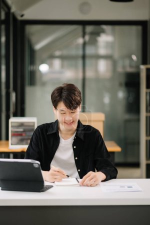 Téléchargez les photos : Jeune homme asiatique attrayant travaillant avec ses notes dans un bureau moderne - en image libre de droit