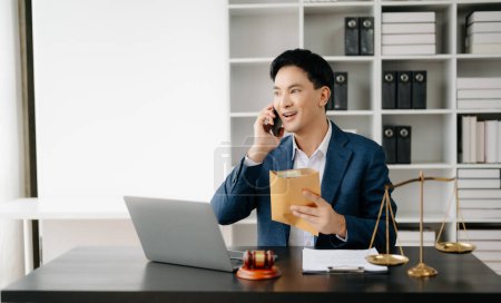 Photo for Asian male lawyer chatting online with client on smartphone, discussing details of lawsuit in modern office - Royalty Free Image