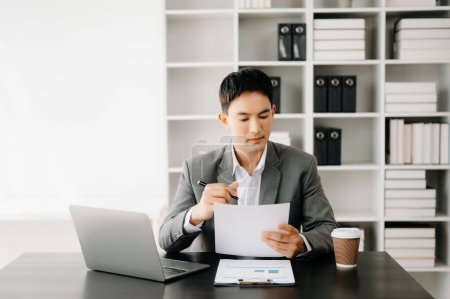 Téléchargez les photos : Jeune homme d'affaires asiatique travaillant au bureau moderne avec ordinateur portable, tablette et prendre des notes sur le papier - en image libre de droit
