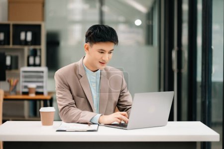 Téléchargez les photos : Jeune homme d'affaires asiatique travaillant avec un ordinateur portable et prenant des notes dans un bureau moderne - en image libre de droit