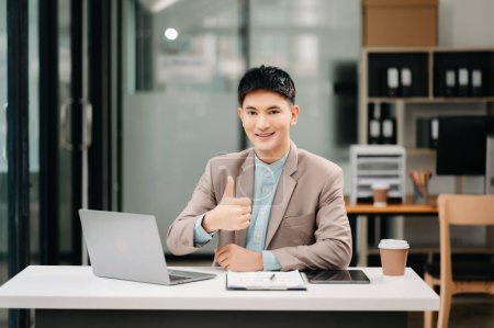 Photo for Young Asian businessman working with laptop and taking notes in modern office, showing thumb up gesture - Royalty Free Image