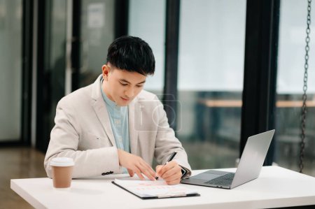 Foto de Joven hombre de negocios asiático trabajando con el ordenador portátil y tomando notas en la oficina moderna - Imagen libre de derechos
