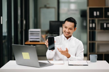 Photo for Smiling Asian businessman showing smartphone in modern office - Royalty Free Image