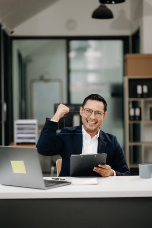 Téléchargez les photos : Heureux homme d'affaires asiatique travaillant avec ordinateur portable et documents dans le bureau moderne. Jeune homme célébrant le succès - en image libre de droit