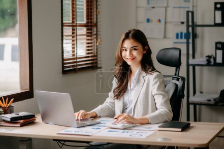 Photo for Young attractive female office worker working on laptop and smiling at camera in office - Royalty Free Image