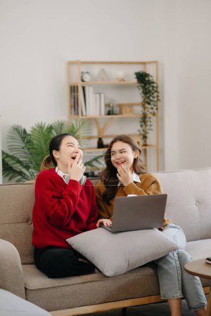 Téléchargez les photos : Deux beauté asiatique souriant jeunes femmes assises sur le canapé, avoir du plaisir à regarder la vidéo de la comédie à la télévision dans la maison. Concept de style de vie d'activité dans le salon - en image libre de droit