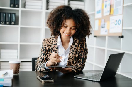 Photo for Young African woman with afro hair, wearing leopard jacket while working on office and using tablet at table with calculator and laptop - Royalty Free Image