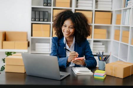 Photo for Startup small business SME. Entrepreneur African woman using laptop to receive and check online orders. Young female small business owner packing parcels to ship them to customers - Royalty Free Image