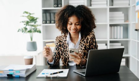 Téléchargez les photos : Jeune femme africaine avec des cheveux afro, portant une veste léopard tout en travaillant sur le bureau et tenant un téléphone mobile - en image libre de droit