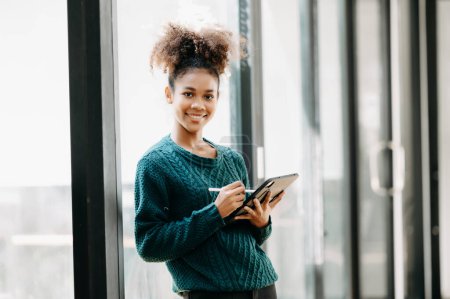 Téléchargez les photos : Heureuse jeune étudiante africaine étudiant en ligne dans la bibliothèque du collège moderne - en image libre de droit