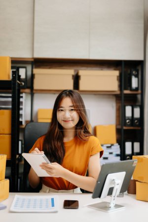 Téléchargez les photos : Petite entreprise en démarrage PME, femme entrepreneur asiatique vérifiant les commandes en ligne et préparant des colis pour l'expédition - en image libre de droit