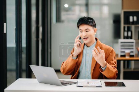 Téléchargez les photos : Souriant asiatique homme d'affaires parler au téléphone et en utilisant un ordinateur portable au bureau - en image libre de droit