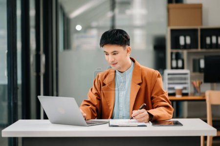Téléchargez les photos : Jeune homme d'affaires asiatique utilisant un ordinateur portable dans un bureau moderne. Souriant confiant cols blancs - en image libre de droit
