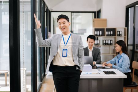 Foto de Joven atractivo asiático hombre de negocios sonriendo en la oficina moderna, sus colegas trabajando en segundo plano - Imagen libre de derechos