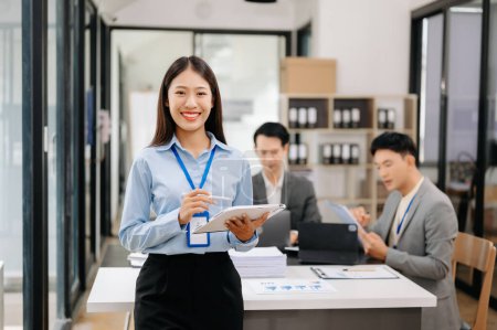 Foto de Joven atractiva mujer de negocios asiática sonriendo a la cámara en la oficina moderna, sus colegas trabajando en segundo plano - Imagen libre de derechos