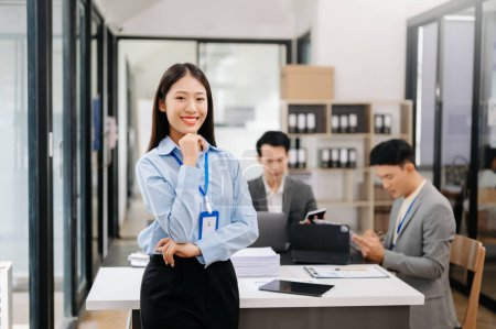Photo for Young attractive Asian businesswoman smiling at camera in modern office, her colleagues working in background - Royalty Free Image