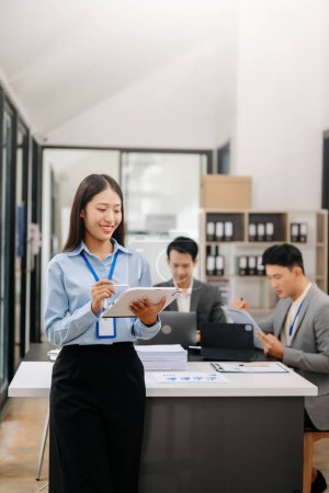 Photo for Young attractive Asian businesswoman smiling at camera in modern office, her colleagues working in background - Royalty Free Image