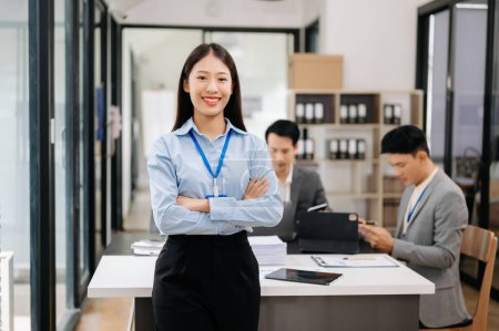 Foto de Joven atractiva mujer de negocios asiática sonriendo a la cámara en la oficina moderna, sus colegas trabajando en segundo plano - Imagen libre de derechos
