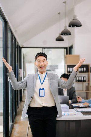 Photo for Young attractive Asian businessman smiling in modern office, his colleagues working in background - Royalty Free Image