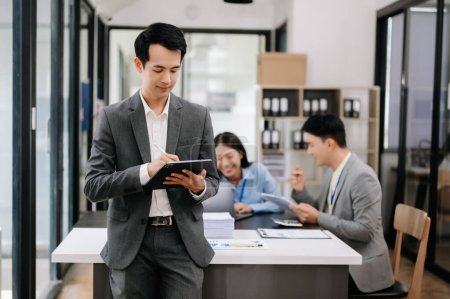 Téléchargez les photos : Jeune homme d'affaires asiatique attrayant souriant dans un bureau moderne, ses collègues travaillant en arrière-plan - en image libre de droit