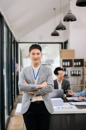 Téléchargez les photos : Jeune homme d'affaires asiatique attrayant souriant dans un bureau moderne, ses collègues travaillant en arrière-plan - en image libre de droit