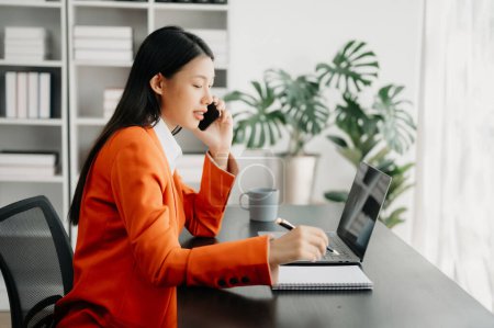 Téléchargez les photos : Jeune attrayante asiatique femme de bureau costume d'entreprise de travail avec bloc-notes et ordinateur portable dans le bureau moderne - en image libre de droit
