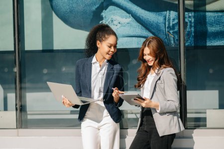 Téléchargez les photos : Femmes d'affaires vont dans le centre-ville dans le style d'affaires décontracté intelligent, parler, travailler ensemble sur tablette numérique - en image libre de droit