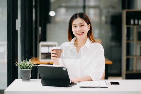 Photo for Young beautiful Asian woman typing on tablet while sitting at the table in office - Royalty Free Image