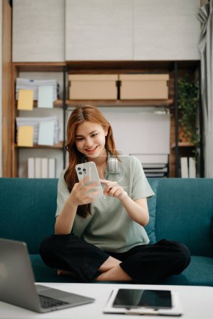Photo for Female student sitting at sofa using smartphone to search an online informations in living room - Royalty Free Image