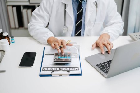 Foto de Concepto de costos y tarifas de atención médica. Doctor usando calculadora y teléfono inteligente en su oficina - Imagen libre de derechos