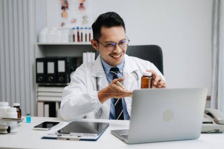 Téléchargez les photos : Confiant jeune médecin masculin en uniforme médical blanc travaillant sur ordinateur - en image libre de droit