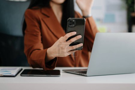 Téléchargez les photos : Femme asiatique confiante avec smartphone et ordinateur portable travaillant dans le bureau. - en image libre de droit