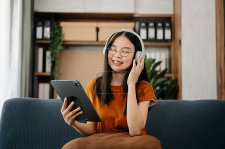 Photo for Smiling Asian girl relaxing at home, she is playing music using digital tablet and wearing white headphones on sofa - Royalty Free Image