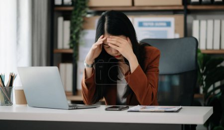 Foto de Mujer asiática sintiendo migraña en la cabeza. Excesiva mujer de negocios financiera trabajando en el ordenador portátil en la oficina - Imagen libre de derechos