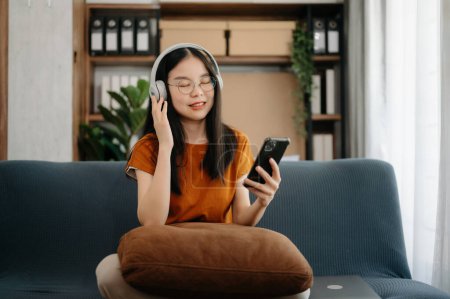 Foto de Sonriente chica asiática que se relaja en casa, está tocando música usando un teléfono inteligente y usando auriculares blancos en el sofá - Imagen libre de derechos